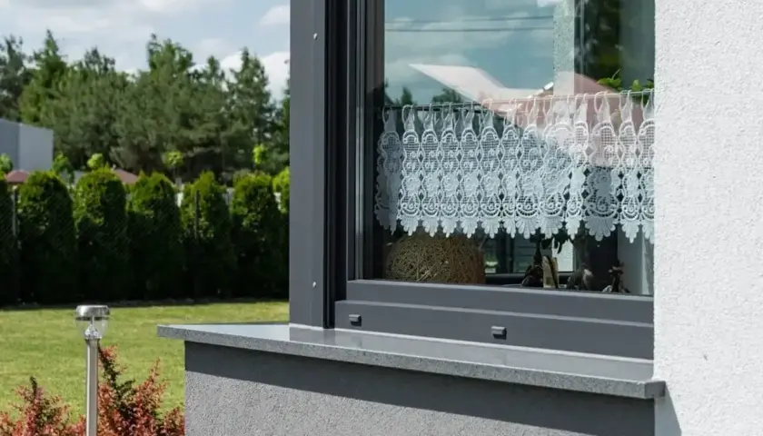 Szary parapet okienny z kamienia, z ozdobną koronkową firanką wewnątrz. Za oknem zielony ogród.