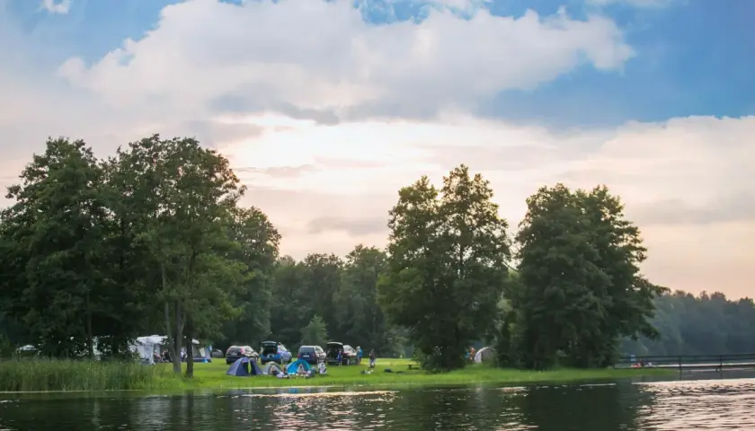 Malowniczy pole campingowe nad jeziorem, z namiotami, samochodami i ludźmi odpoczywającymi wśród drzew.