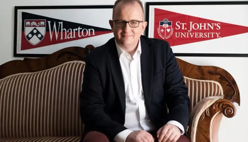 Mężczyzna w garniturze siedzi na kanapie. W tle banery Wharton i St. John's University, prestiżowa uczelnia w USA.