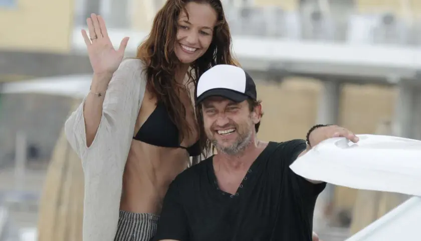 Gerard Butler z uśmiechniętą kobietą, która może być jego żoną, na plaży. Ona macha ręką, on ma na głowie czapkę.