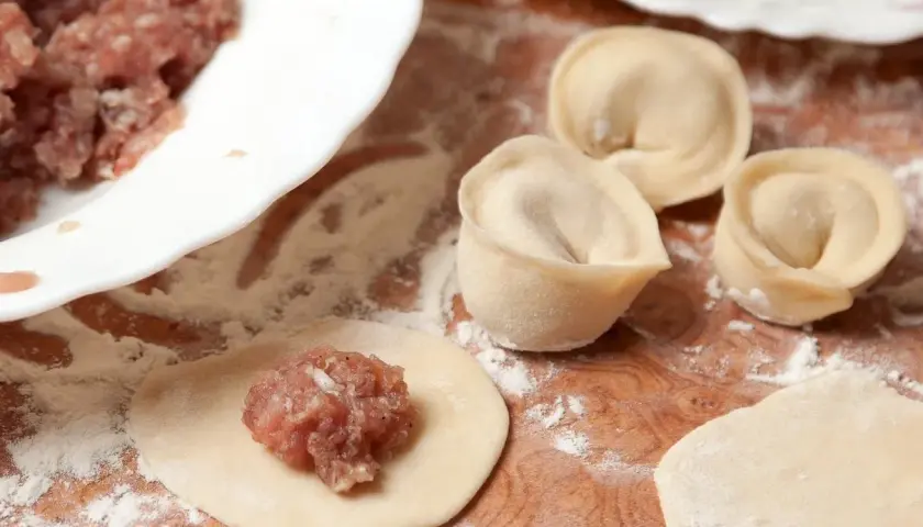 Przygotowanie pierogów z mięsem mielonym surowym. Na drewnianej desce leżą surowe pierogi, obok miska z farszem.