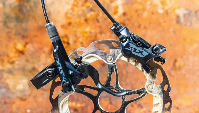 Shimano SLX M7120 hydraulic disc brakes, ready for any trail. The dark blue calipers and rotors are mounted on a rusty, textured background.