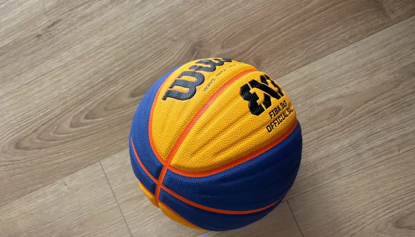 Piłka do koszykówki 3x3 Wilson z pomarańczowo-niebieskimi panelami leży na drewnianej podłodze.