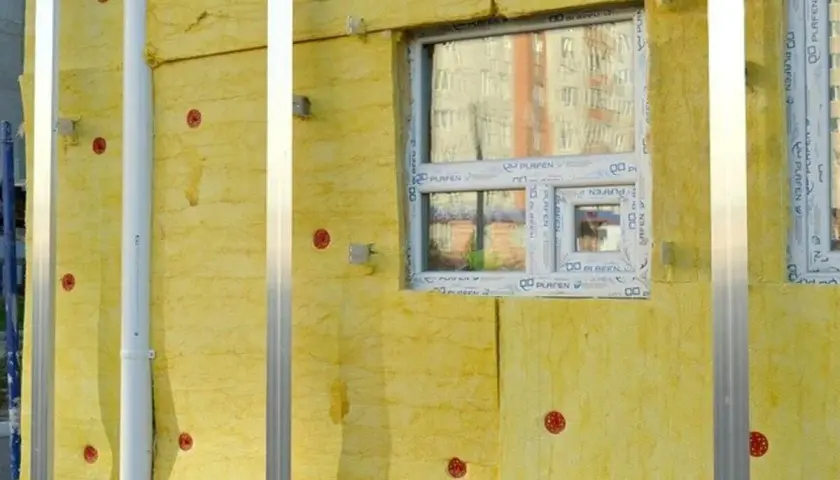 Isolation jaune sur un mur, avec des fenêtres et des fixations rouges. Un conduit blanc traverse l'isolant, assurant un **rupteur thermique** efficace.