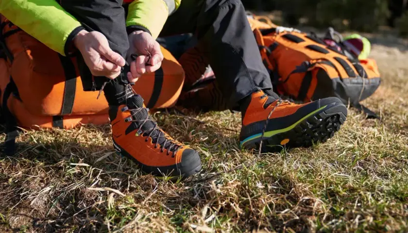 A person is lacing up bright orange hiking boots with a heel lock lacing system, preparing for an adventure with an orange backpack nearby.
