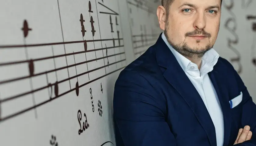 Mężczyzna w granatowej marynarce, oparty o ścianę z nutami. Wygląda jak człowiek do wszystkiego, gotowy na każde wyzwanie.