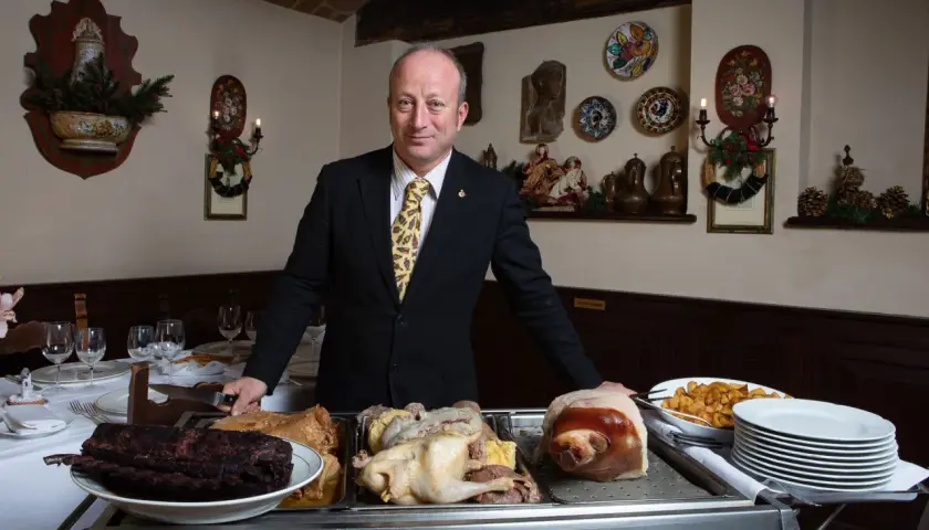 A man in a suit stands behind a cart with various meats, including ribs, a whole chicken, and a pig's head.