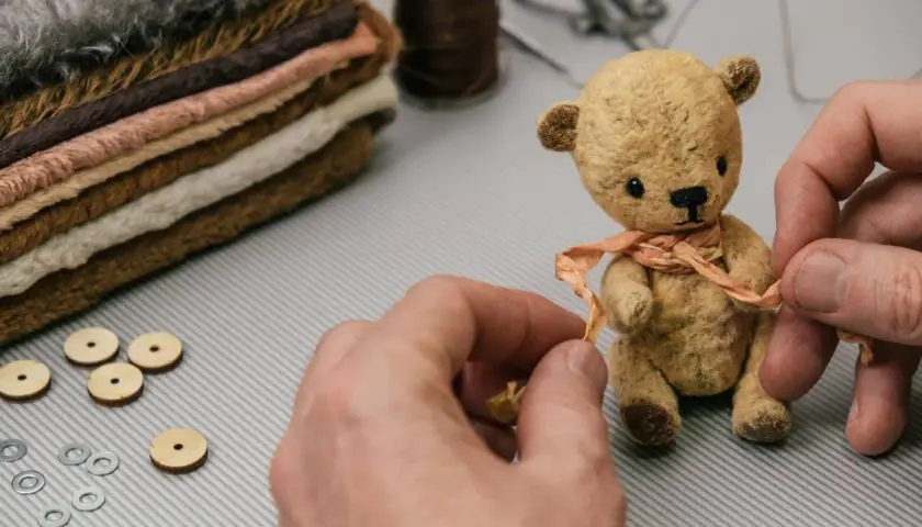 Mały, ręcznie robiony miś z materiału, ozdabiany wstążką, obok stosu tkanin i akcesoriów.