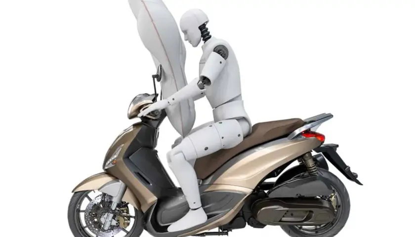 Un maniquí en una moto dorada con un airbag inflado protegiendo su torso. Las motos tienen airbag para mayor seguridad.