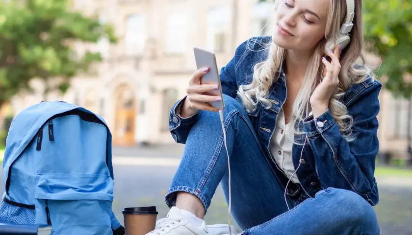 Dziewczyna w dżinsowej kurtce słucha muzyki na telefonie. Obok niej leży niebieski plecak i książki. Idealny moment, by pomyśleć, o czym można pisać z chłopakiem.