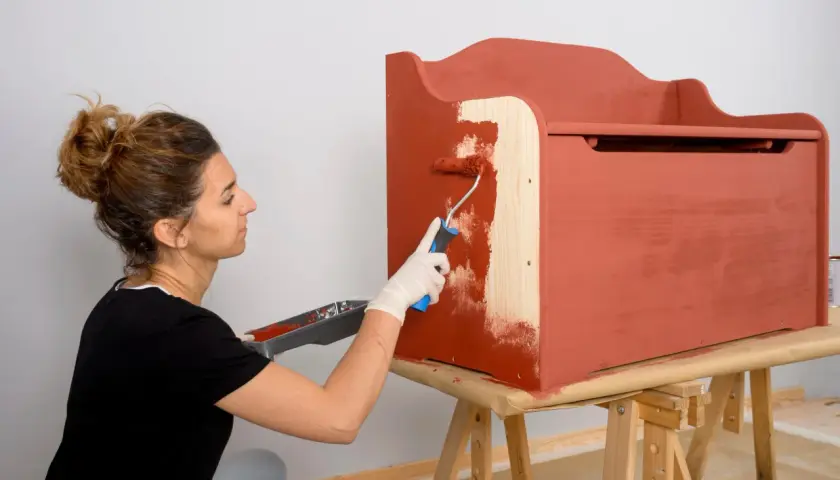 Mujer con guantes blancos pinta con rodillo una caja de madera de color terracota.