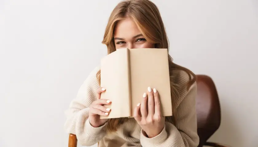Dziewczyna z uśmiechem ukrywa twarz za książką, zanurzona w świecie literatury young adult.
