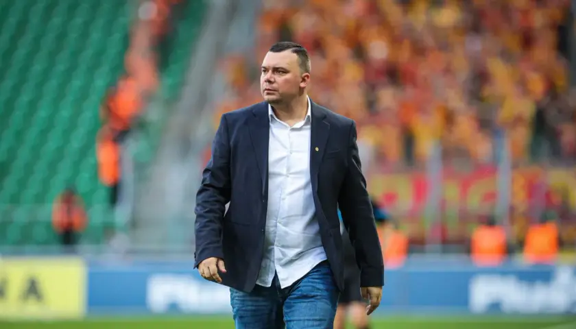 Trener Jagiellonii w eleganckim garniturze i dżinsach, na tle trybun stadionu wypełnionych kibicami.