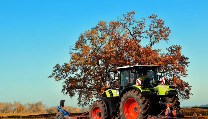 Zielony traktor z pługiem pod jesiennym drzewem. Może posłużyć jako inspiracja do wypełnienia wniosku o odrolnienie działki wzór.