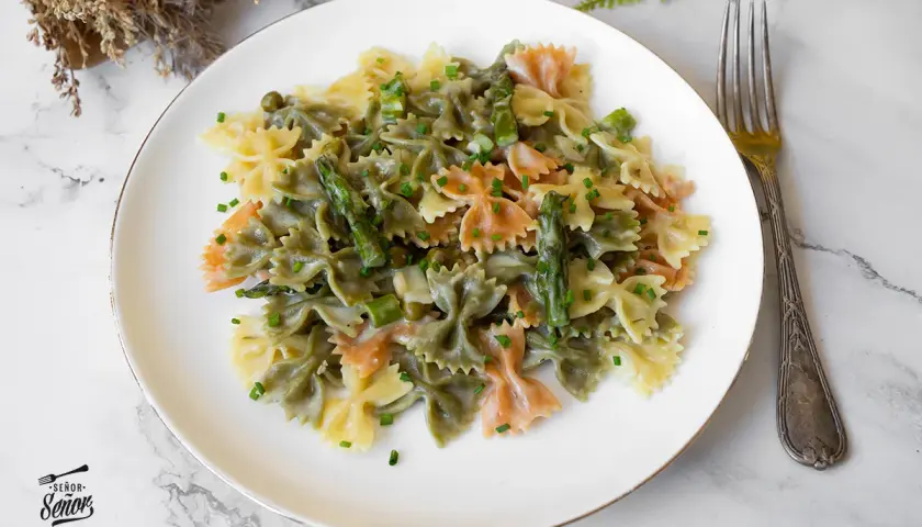 Plato de pasta primavera con farfalle tricolor, espárragos y guisantes en salsa cremosa, adornado con cebollino picado.