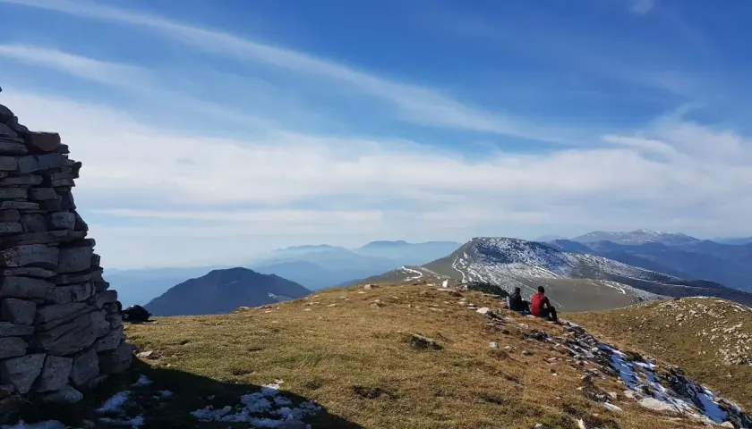 Dos personas contemplan el paisaje montañoso desde el Puig d'Estela, con un muro de piedra a la izquierda y el cielo azul con nubes.
