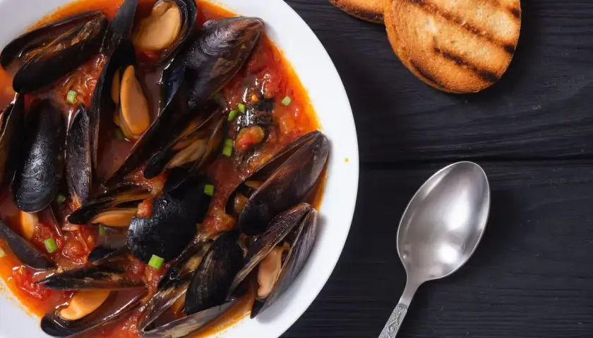 Un invitante piatto di zuppa di cozze in un ricco sugo di pomodoro, guarnito con erba cipollina, accompagnato da crostini tostati e un cucchiaio.