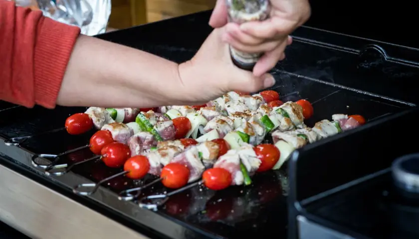 Brochettes de viande, tomates et courgettes sur une plancha à gaz. Une main assaisonne les brochettes.