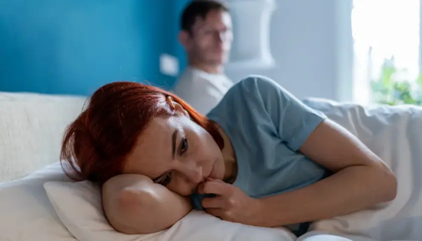 Smutna kobieta leży w łóżku, czując się samotna i przygnębiona. Jej partner jest w tle, ale nie okazuje wsparcia, co potęguje poczucie poniżania w związku.