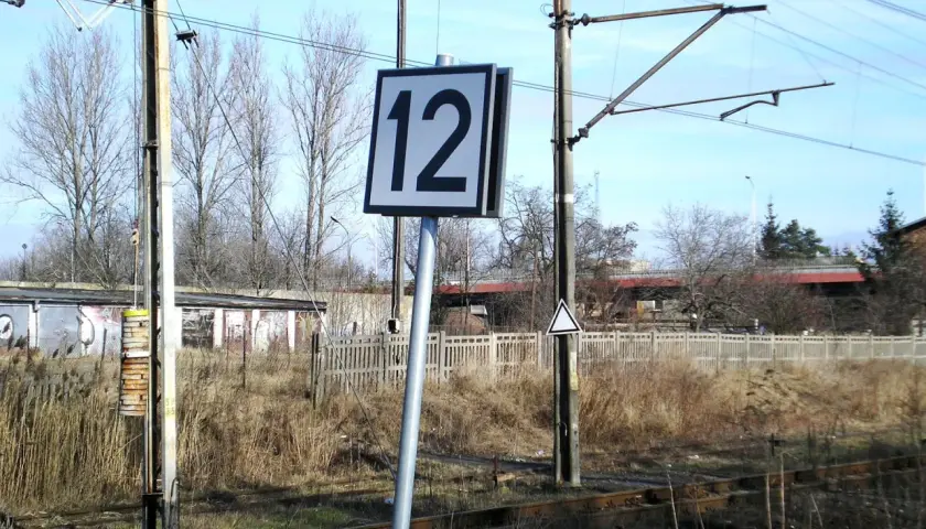 Wskaźnik W24 z numerem 12 przy torach kolejowych. W tle budynki i drzewa.