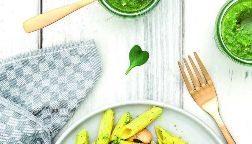 Makaron penne z zielonym pesto, podany na talerzu, obok słoików z pesto i złotego widelca.