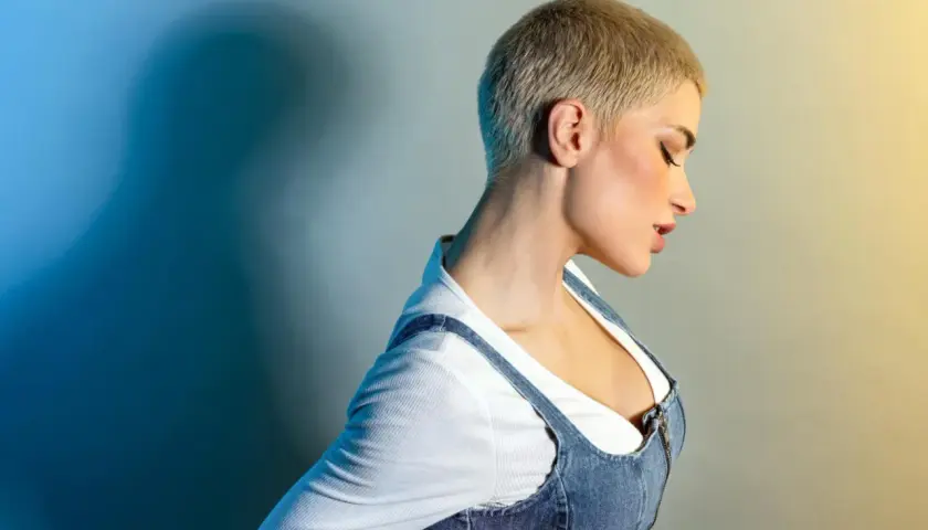 Kobieta z fryzurą buzz cut blond, w białej bluzce i dżinsowej sukience na szelkach, z profilu, z zamkniętymi oczami.