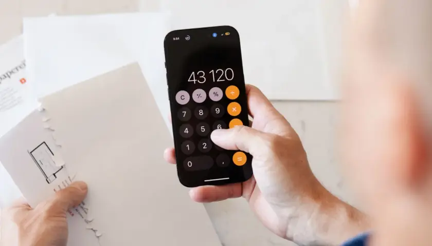Dłoń z telefonem wyświetlającym kalkulator, na którym widnieje kwota 43120. Obok leżą białe koperty, sugerujące problemy z zadłużeniem.
