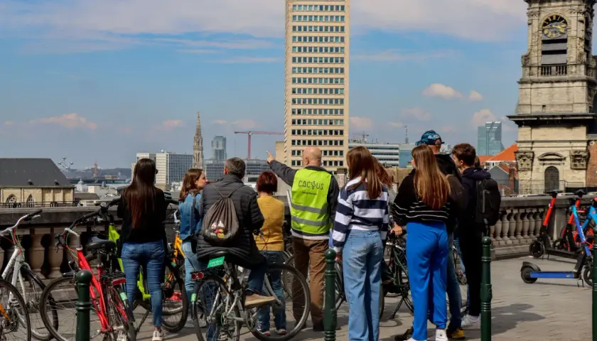 A group gathers for Brussels bike tours, with a guide pointing towards the city skyline featuring a tall skyscraper and an old clock tower.