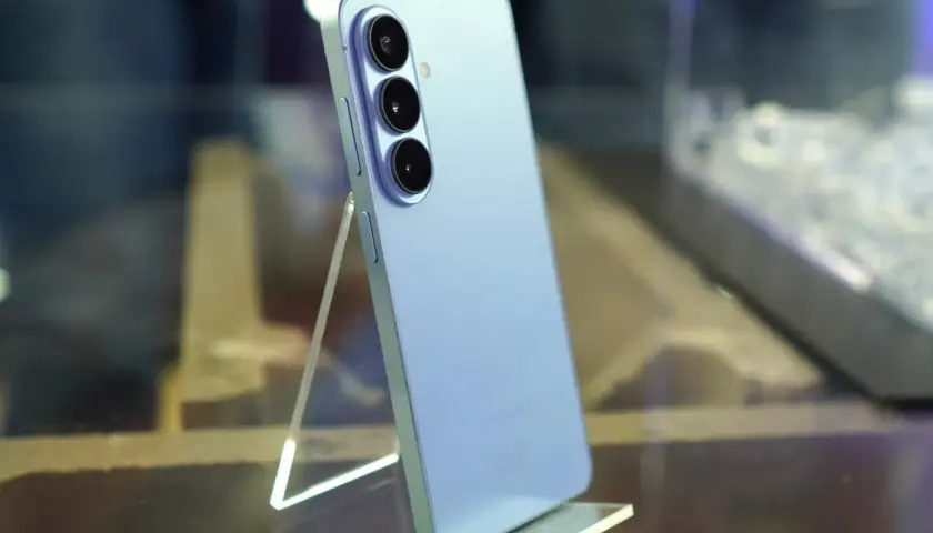 Jasnoniebieski Samsung Galaxy S26 z potrójnym aparatem z tyłu, stojący na przezroczystej podstawce.