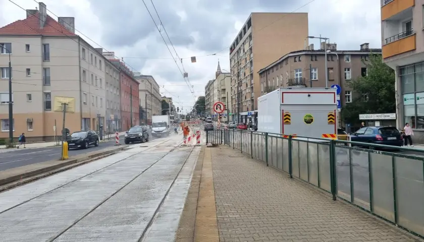 Ulica z torami tramwajowymi w remoncie, budynki mieszkalne, samochody i pracownicy w pomarańczowych kamizelkach.