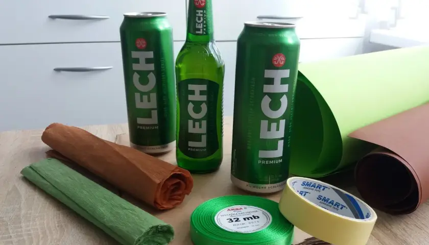 Puszki i butelka piwa Lech, rolki papieru, wstążka i taśma klejąca na drewnianym stole.