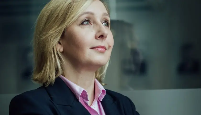 Blond polityk w różowej koszuli i granatowej marynarce patrzy w bok. W tle rozmyte sylwetki innych polityków.