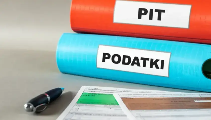Dwa segregator, jeden czerwony z napisem PIT, drugi niebieski z napisem PODATKI, leżą na biurku obok formularzy podatkowych i długopisu. Czas na zwolnienie z PIT!