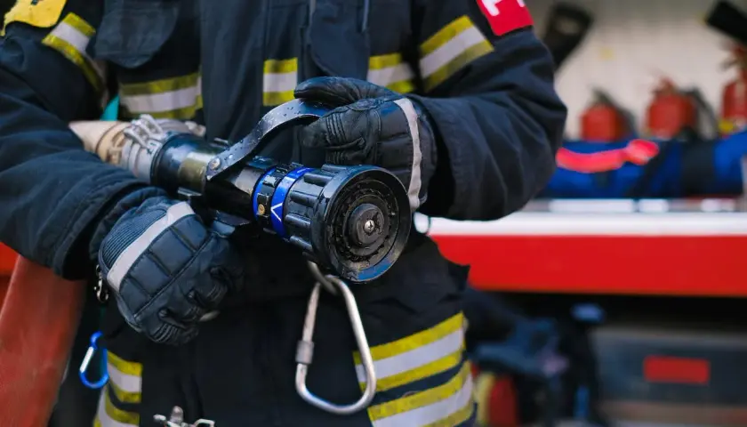 Strażak w rękawicach trzyma prądownicę. Zastanawiasz się, jakie są stopnie w PSP po ilu latach?