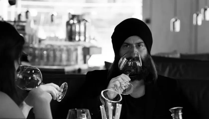 Ein Mann mit Bart und Mütze trinkt Wein. Er hält das Glas am Stiel, eine elegante Art, wie trinkt man wein.