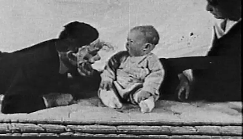 Bambino seduto, un uomo con maschera e una donna sullo sfondo, scena dall'esperimento piccolo Albert.
