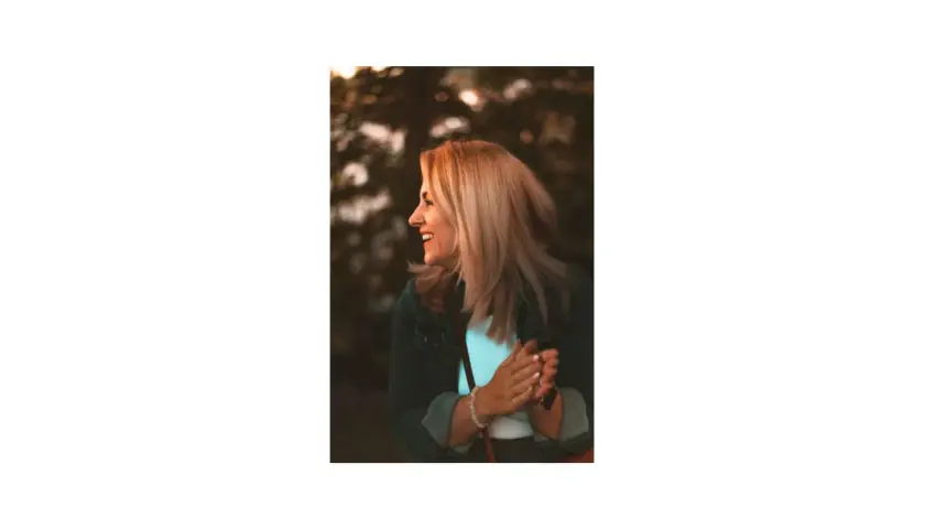 Uśmiechnięta blondynka w dżinsowej kurtce, patrząca w bok, na tle drzew.