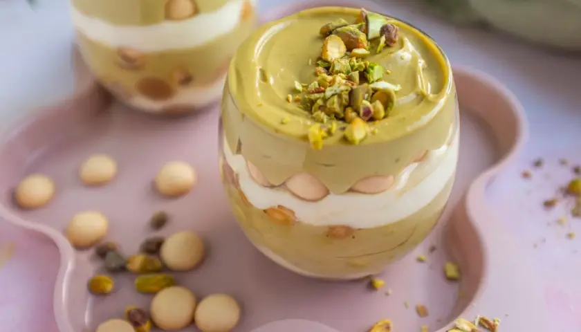 Pyszna tarta pistacjowa w pucharku, przełożona kremem i biszkoptami, posypana orzechami.