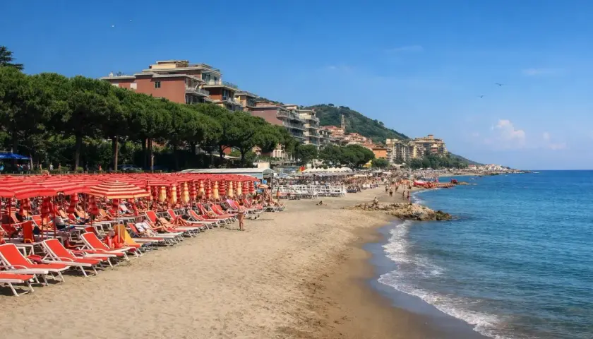 Plage d'Imperia animée, avec des rangées de parasols rouges et des chaises longues sur le sable. La mer bleue étincelle sous un ciel clair.