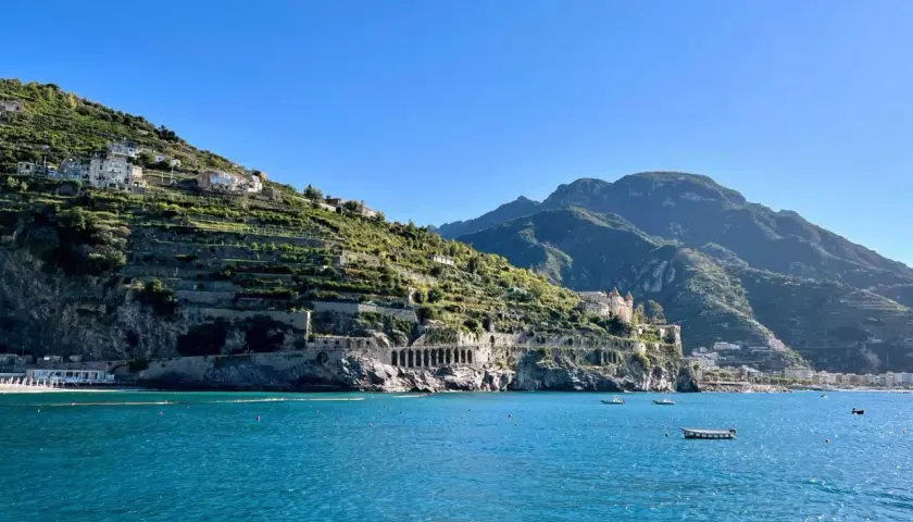A scenic view from a Naples to Amalfi Coast ferry, showing terraced hillsides dotted with houses, a grand villa, and the sparkling blue sea.