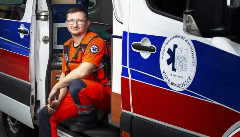 Mężczyzna w pomarańczowym stroju ratownika medycznego siedzi w otwartych drzwiach karetki.