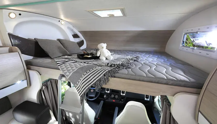 Lit de pavillon de camping-car avec oreillers, couverture et ours en peluche. Vue sur le poste de conduite.