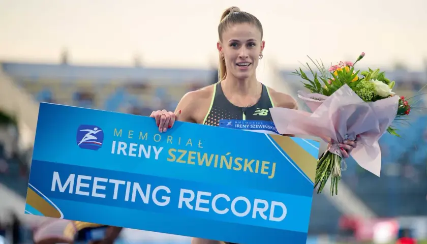 Uśmiechnięta lekkoatletka z bukietem kwiatów trzyma tablicę "MEETING RECORD" z logo memoriału Ireny Szewińskiej.