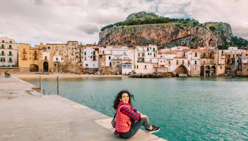 A woman smiles, enjoying her first time visiting Sicily. She sits on a stone pier overlooking the turquoise sea and a picturesque town with a dramatic cliff behind it.