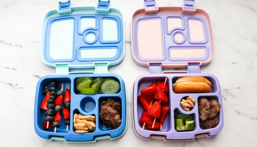 Two bento boxes, one blue and one purple, are filled with lunch items. The blue box contains fruit skewers, cucumber slices, crackers, and ground meat. The purple box holds strawberry skewers, a bun, nuts, and cucumber slices.