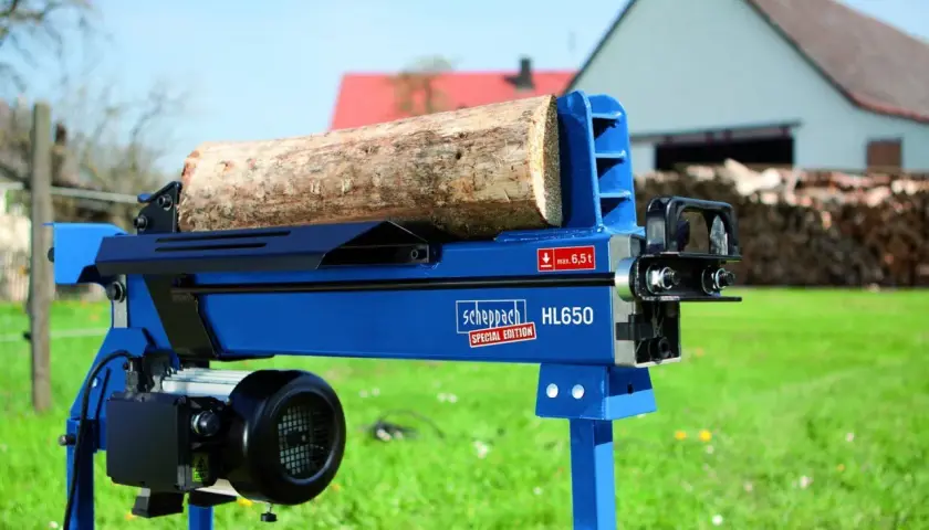 Ein liegender bgu Holzspalter HL650 von Scheppach mit einem Holzstamm bereit zum Spalten. Im Hintergrund ein Haus und Holzstapel.