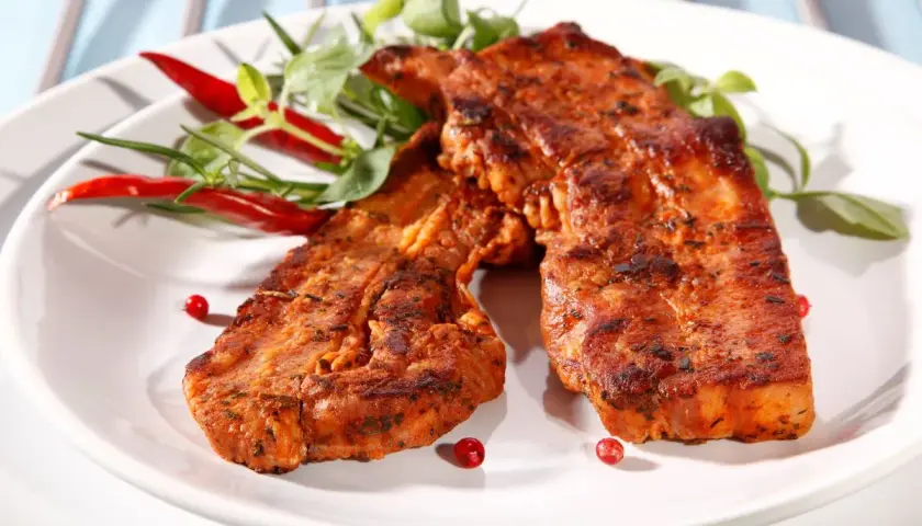 Dwa soczyste plastry boczku na grilla, przyprawione i udekorowane papryczkami chili i zieleniną.