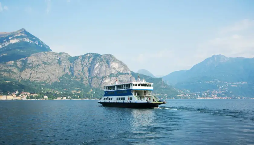 Un ferry transporte des voitures sur le lac de Côme, naviguant paisiblement entre des montagnes majestueuses sous un ciel bleu.