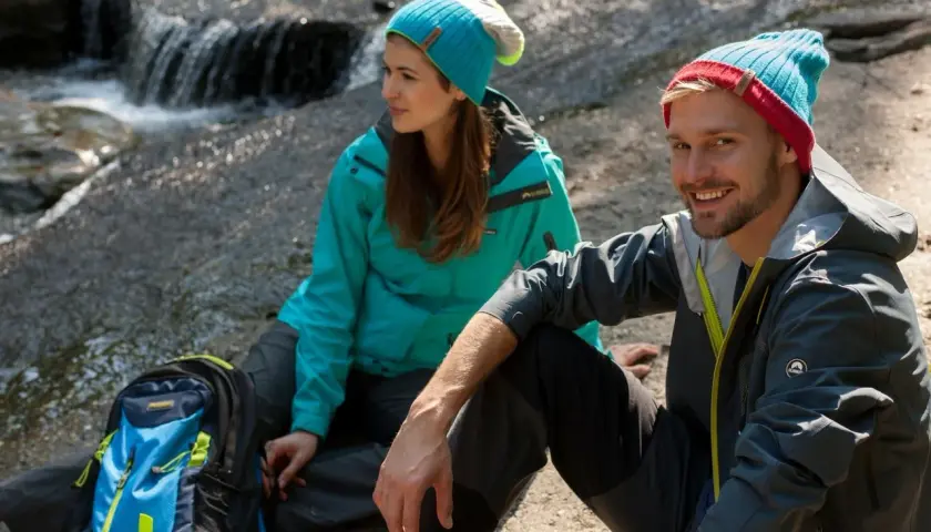 Para, co to jest trekking? Para w czapkach siedzi przy strumieniu, obok plecak.