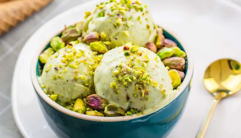Tre palline di cremoso gelato al pistacchio, guarnite con granella e pistacchi interi, in una ciotola blu.